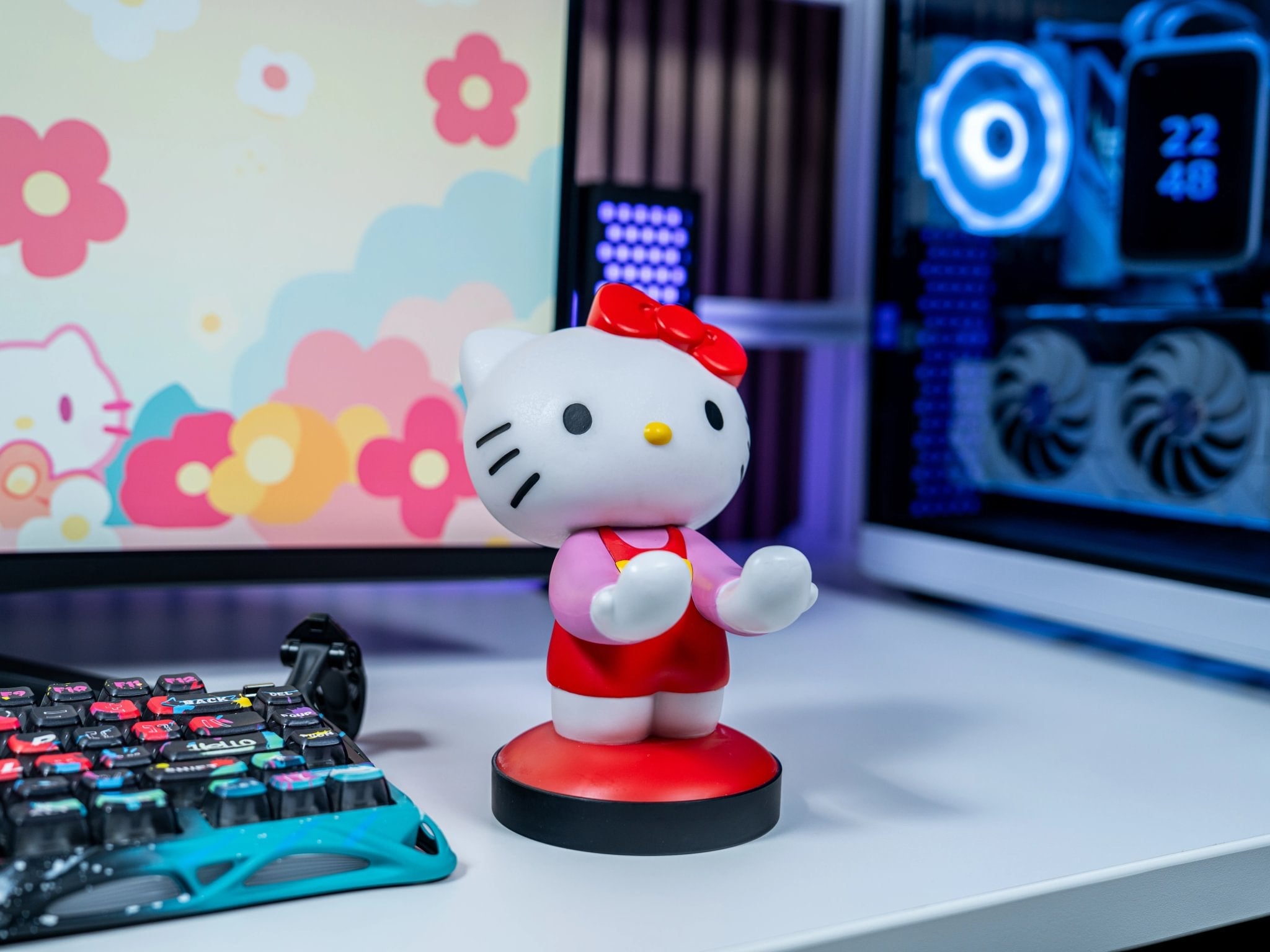 EXG Pro's Hello Kitty Cable Guy controller and phone holder featuring Hello Kitty in her classic red outfit and bow, displayed on a gaming desk with a colorful floral background and PC setup.
