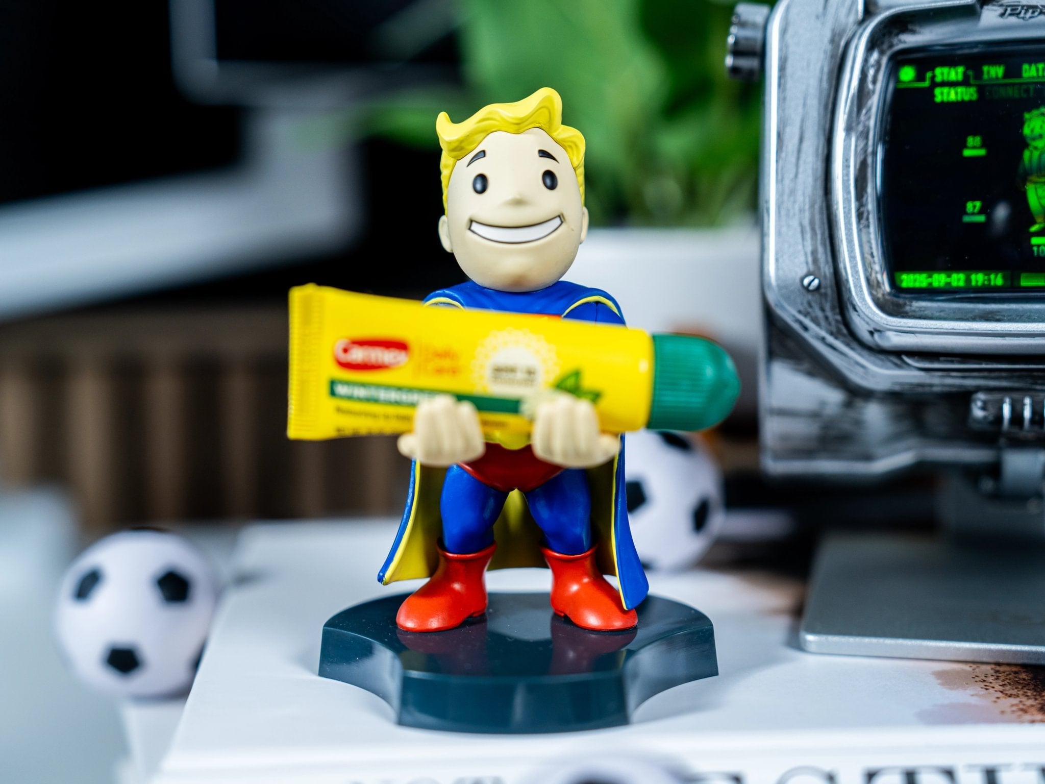 Vault Boy Mini Cable Guy from the Fallout series holding a yellow Carmex lip balm tube, displayed beside a Pip-Boy screen and small soccer ball on a white desk.