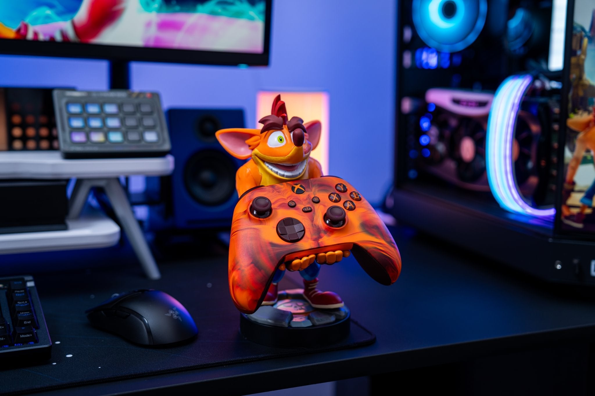 Crash Bandicoot Cable Guy holding an orange Xbox controller on a gaming desk with RGB lighting and monitors.