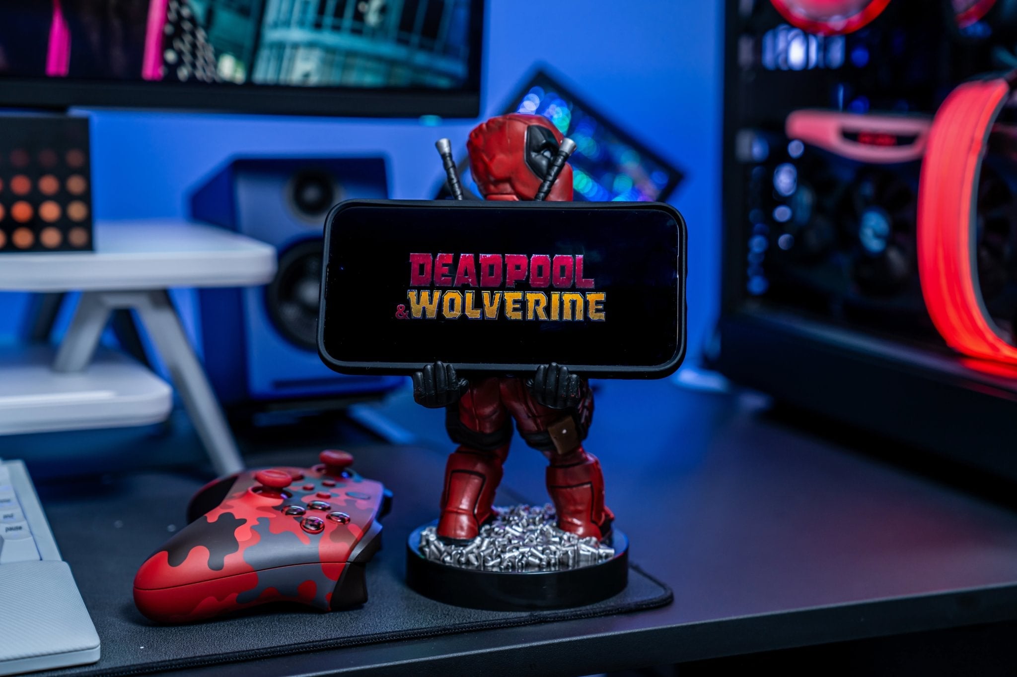 Deadpool Cable Guy holding a smartphone displaying Deadpool & Wolverine on a gaming desk with red and blue RGB lighting.