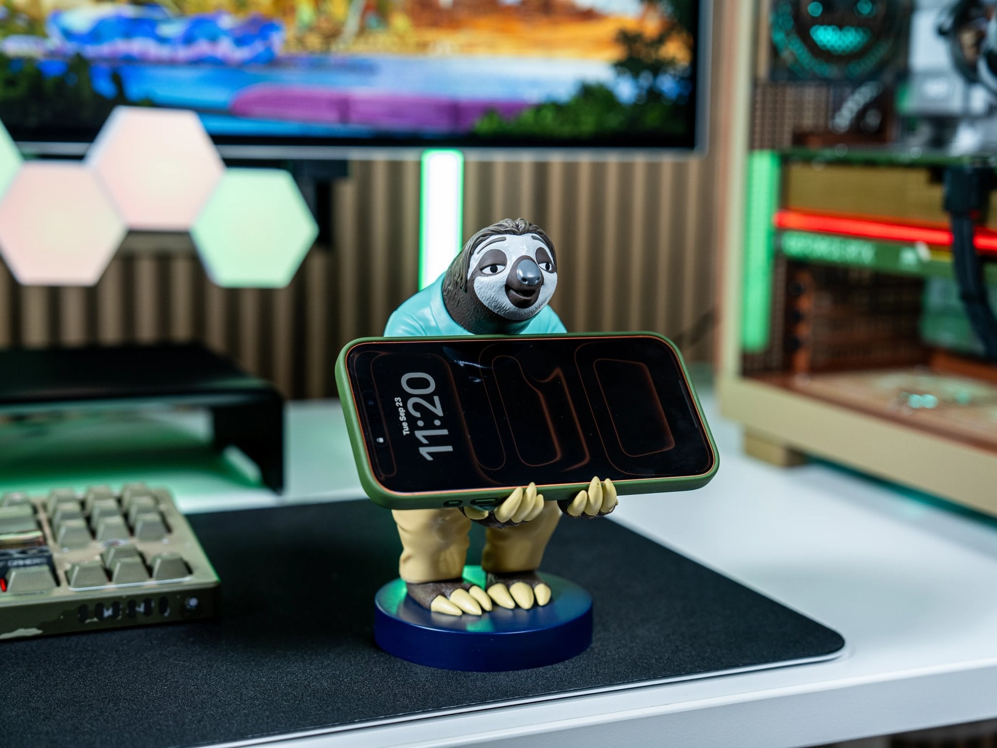 Flash the Sloth Cable Guy collectible from Disney’s Zootopia holding a smartphone on a gaming desk with a modern RGB setup.