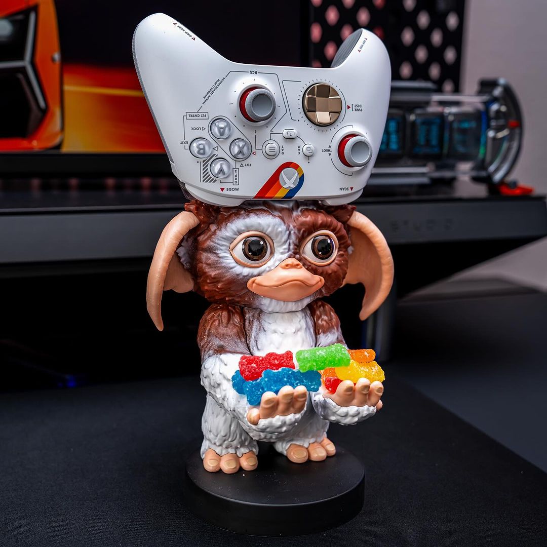 Gizmo Cable Guy from Gremlins holding a colorful candy accessory with a gaming controller balanced on its head, displayed on a modern gaming desk setup.
