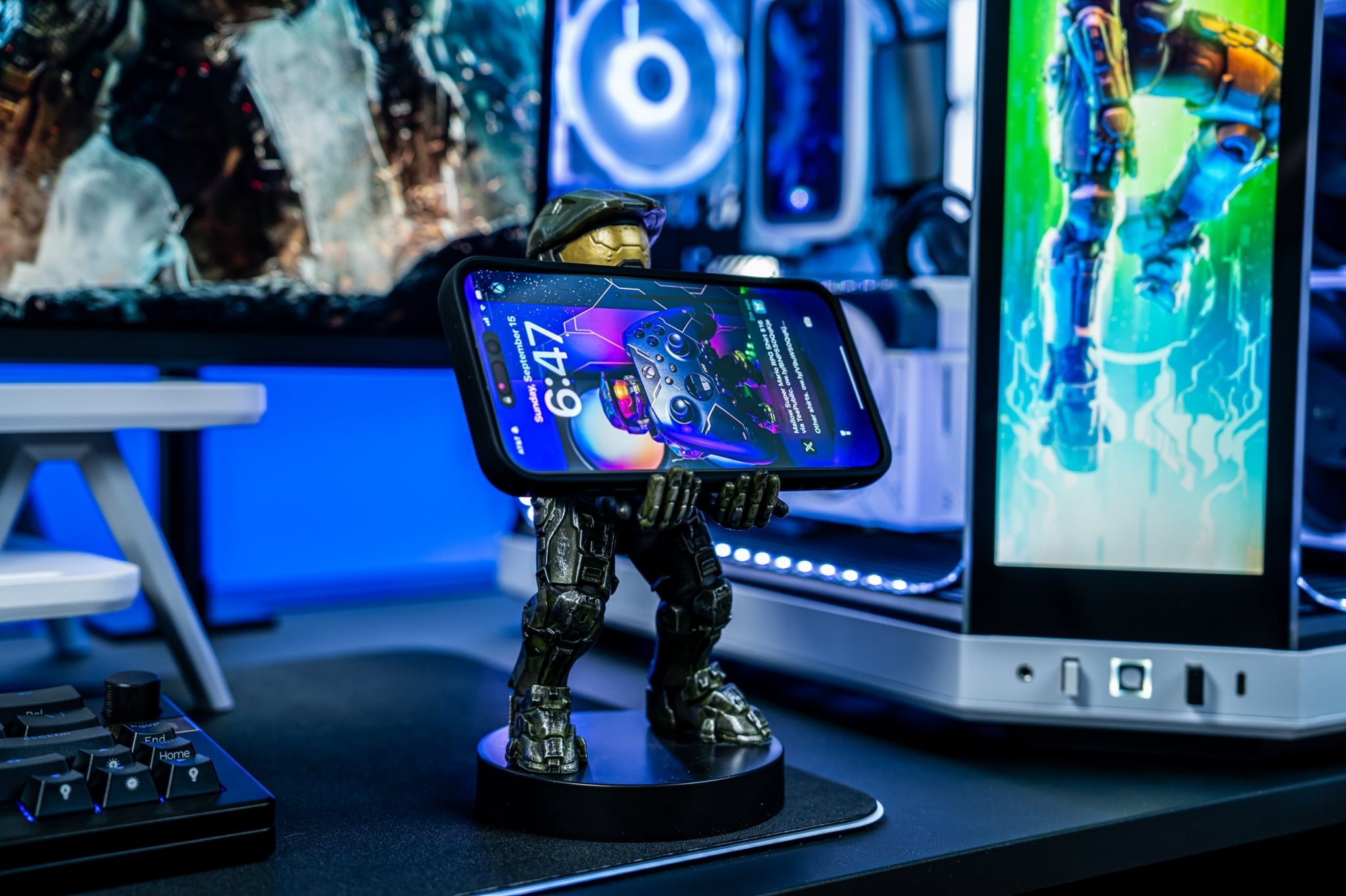Master Chief Cable Guy holding a smartphone on a gaming desk setup, surrounded by illuminated screens and blue RGB lighting.