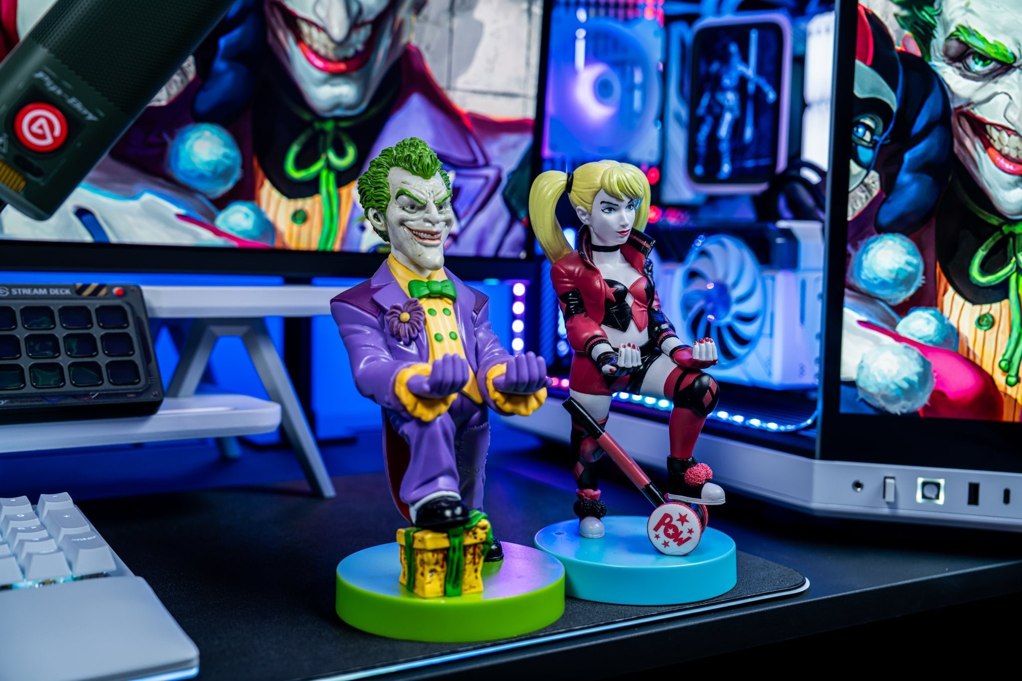 Harley Quinn and The Joker Cable Guys holding devices on a gaming desk with RGB lighting and comic artwork in the background.