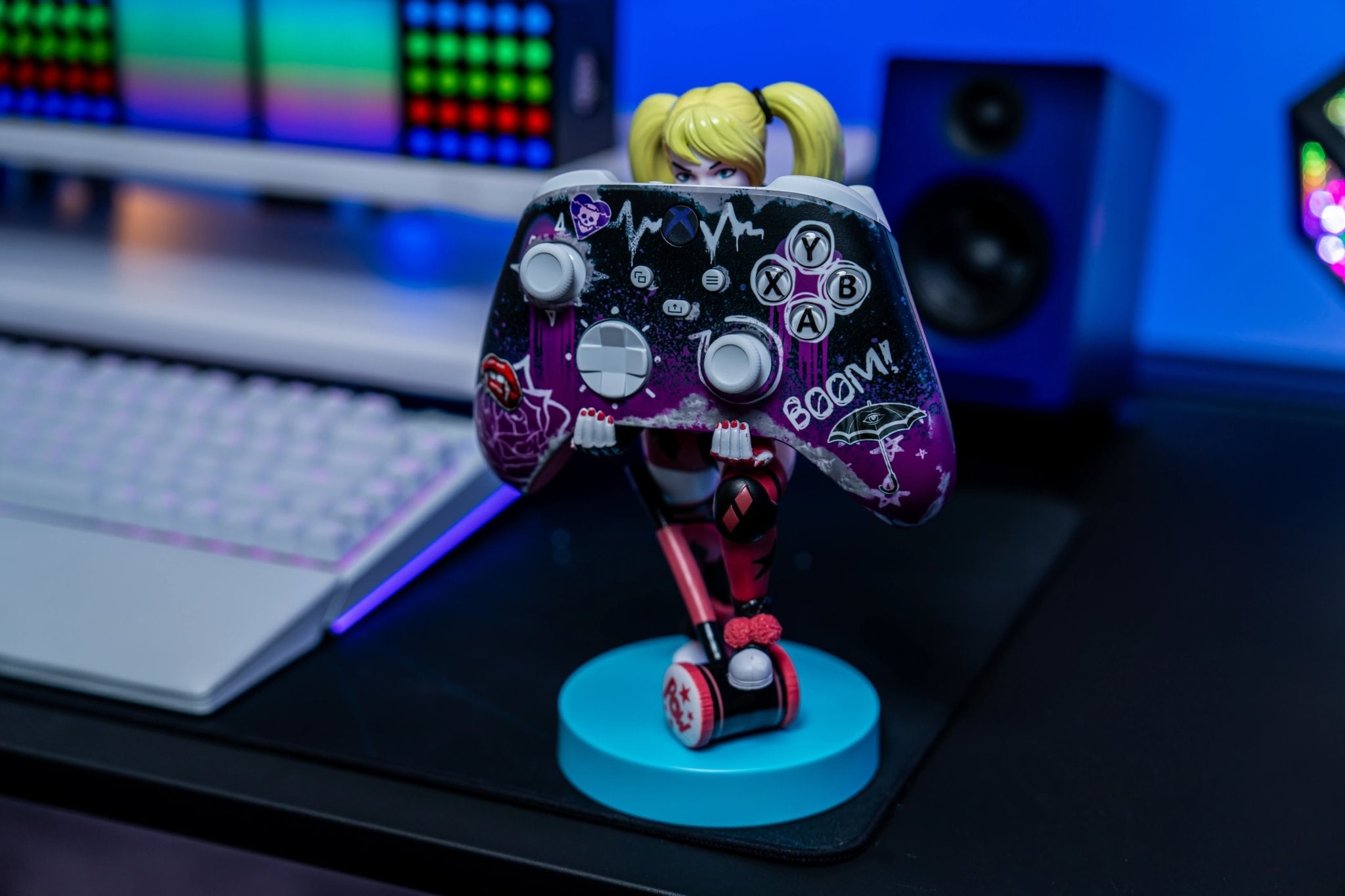 Harley Quinn Cable Guy controller holder standing on a blue base, holding a custom purple and black Xbox controller with graffiti-style art and the word “BOOM!” in a colorful gaming setup.