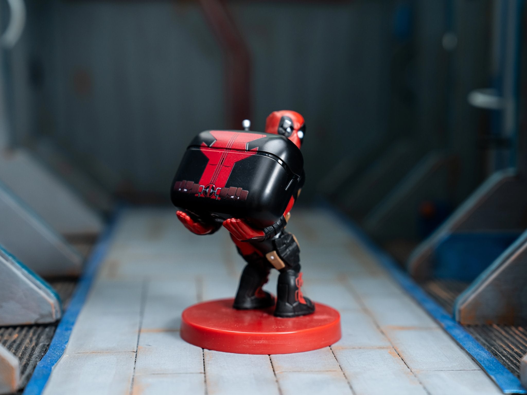 Deadpool Cable Guy holding a matching red and black earbuds case, displayed on a sci-fi themed surface with metallic blue and grey details.