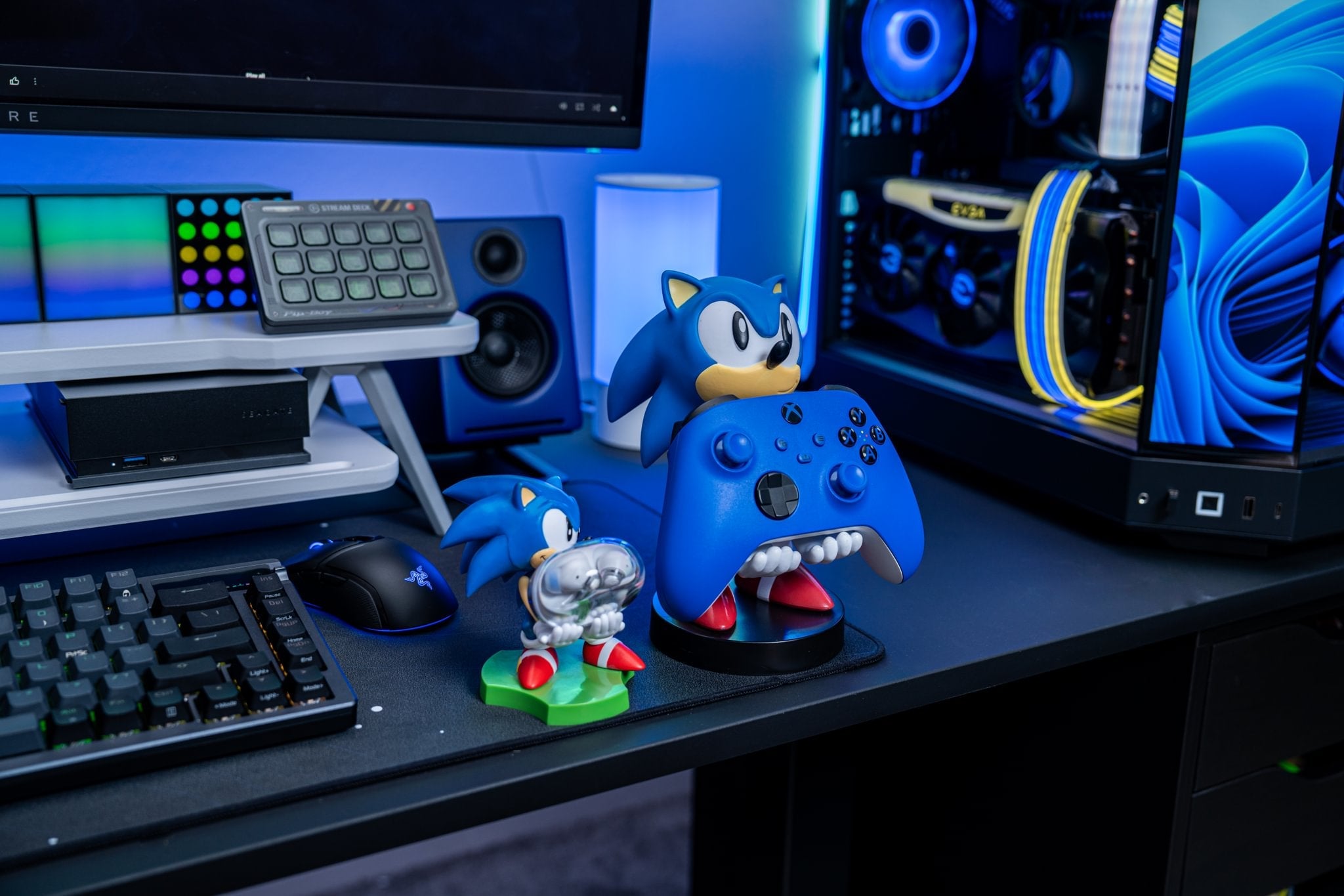 Sonic the Hedgehog Cable Guy and Mini Cable Guy holding an Xbox controller and earbuds on a gaming desk with blue RGB lighting.