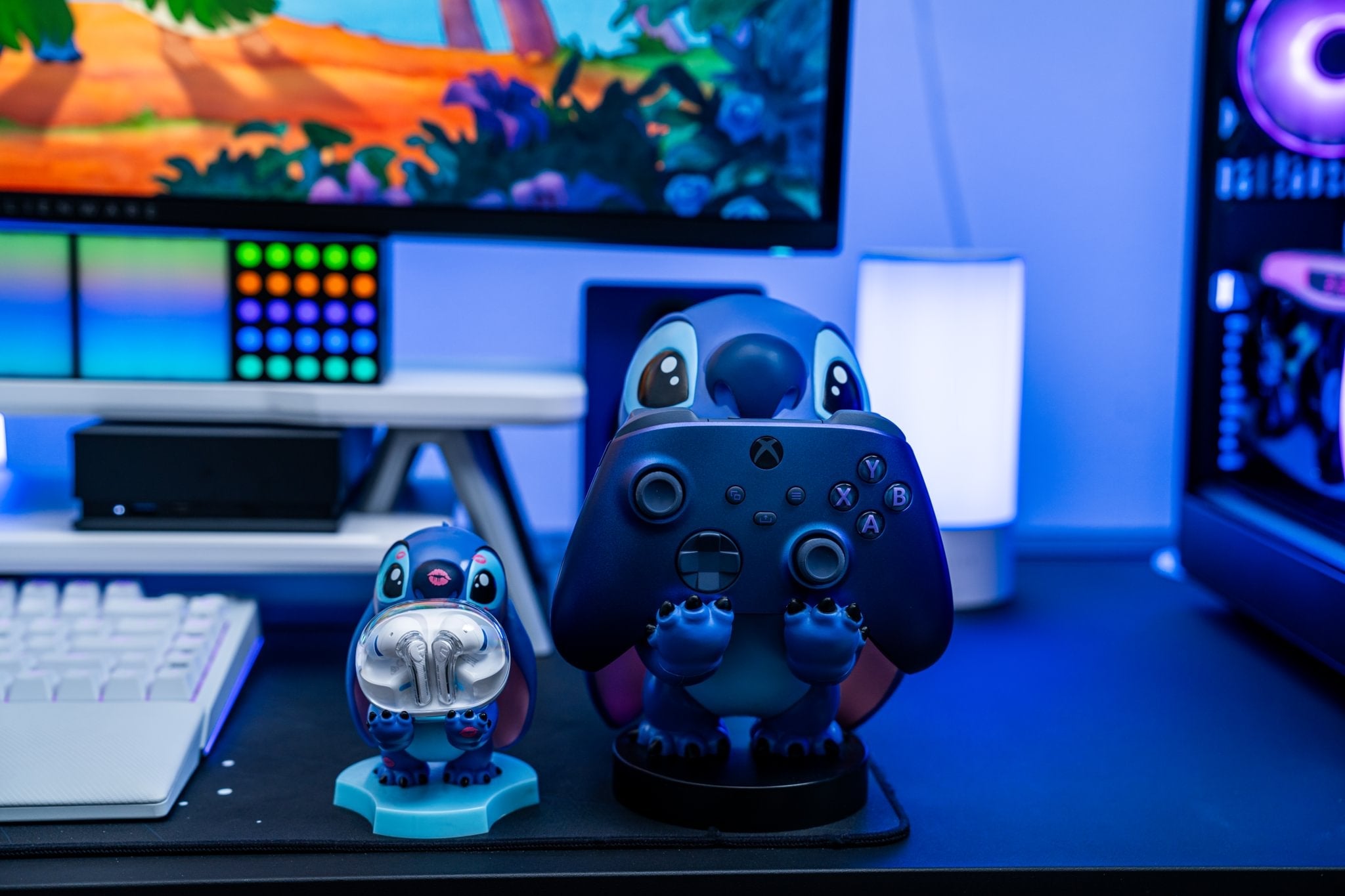 Stitch Cable Guy and Mini Cable Guy figures holding an Xbox controller and earbuds on a gaming desk with a tropical screen background
