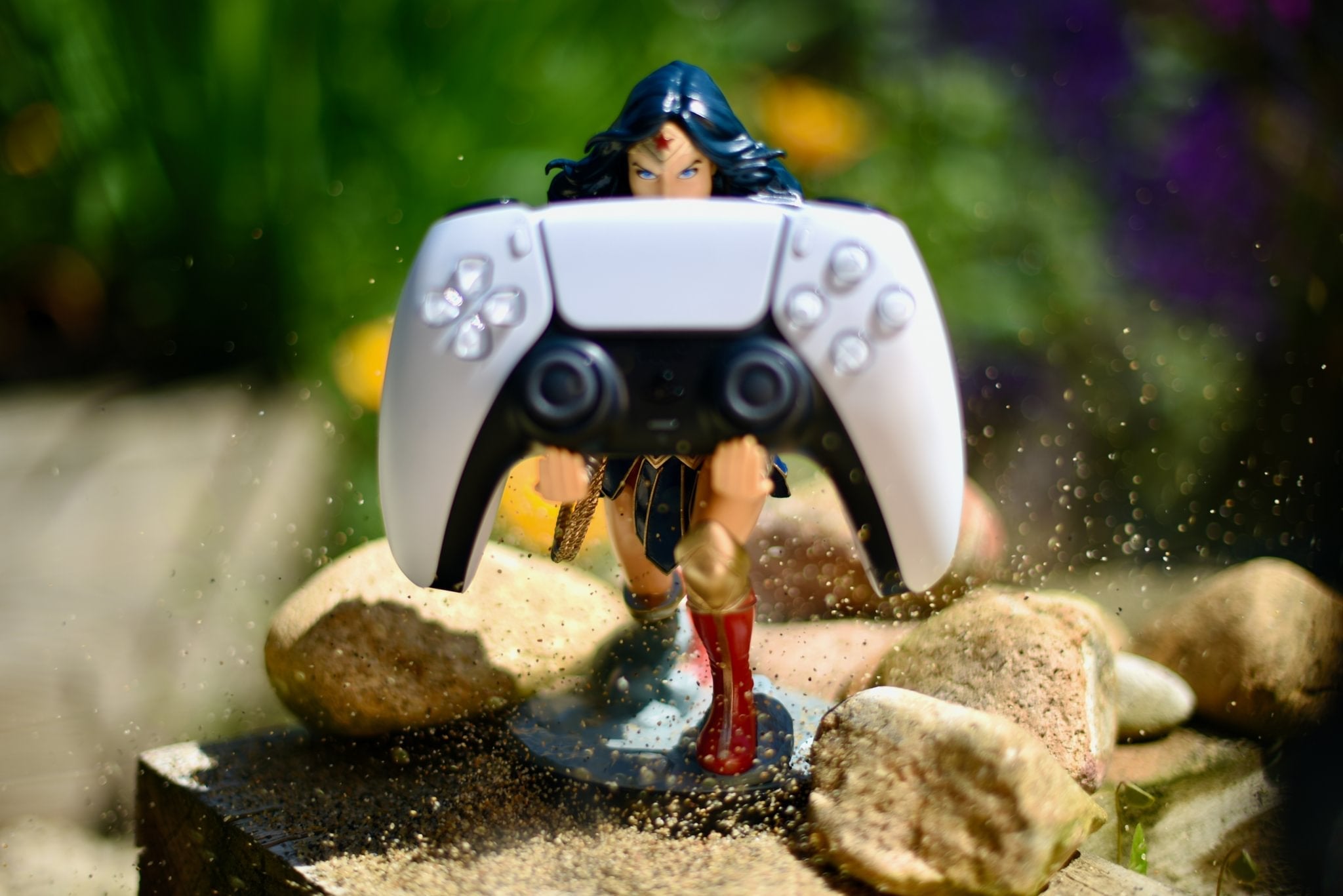 Wonder Woman Cable Guy collectible holding a PlayStation controller outdoors, surrounded by rocks and flying dust for a dynamic action effect.