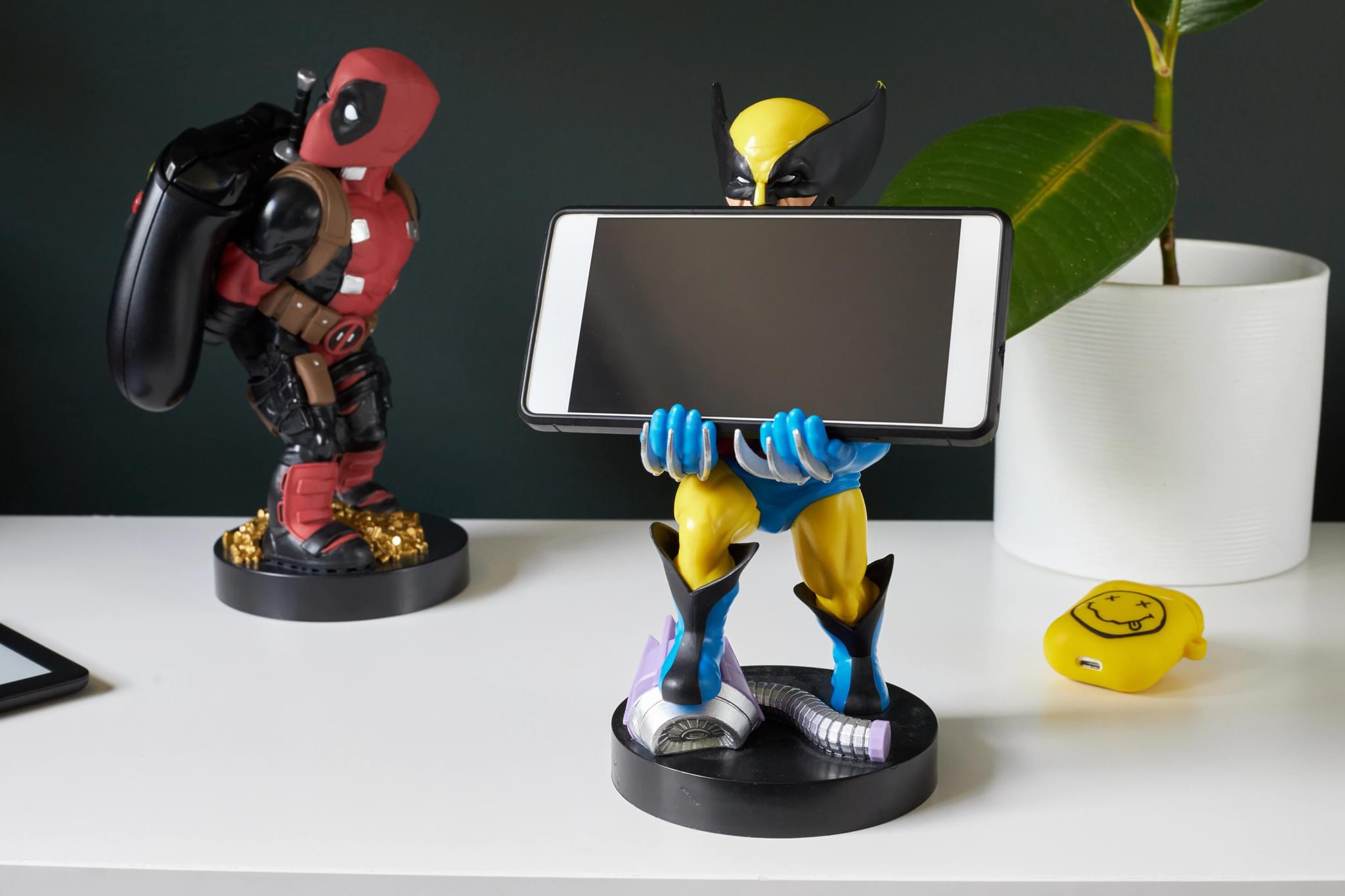Wolverine Cable Guy holding a smartphone with Deadpool Cable Guy in the background, both displayed on a white desk with a plant and accessories nearby.