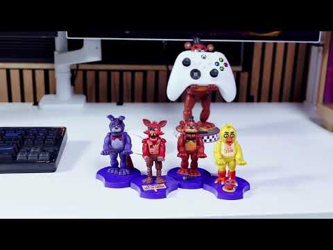 Four stylized Five Nights at Freddy’s Mini Cable Guys figures displayed on blue stands inside a dim, game-themed room. From left to right are small collectible versions of Freddy, Chica, Bonnie, and Foxy, each posed with arms extended as device holders. A small cupcake figure sits in front of Chica. The background includes dark props and an old CRT monitor showing static.