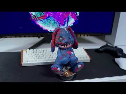 A Call of Duty Mister Peeks Cable Guy figure displayed on a gaming setup, standing upright with arms extended to hold a controller. The character features its signature masked look and tactical styling, set against a lit gaming desk backdrop.