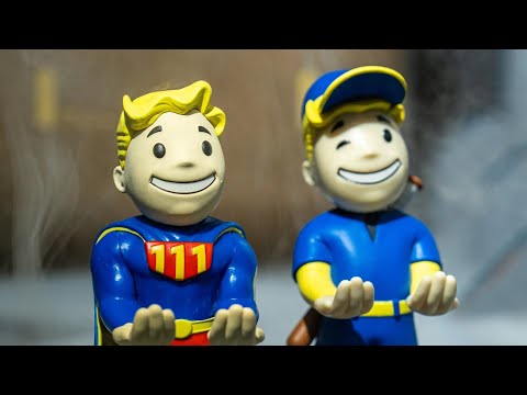 Two Fallout Mini Cable Guys styled as Vault Boy perks: one modeled on the Big League perk holding a baseball bat, and one modeled on the Toughness perk with crossed arms and a confident stance. Both are small collectible holders in Vault-Tec blue and yellow, posed on compact display bases.