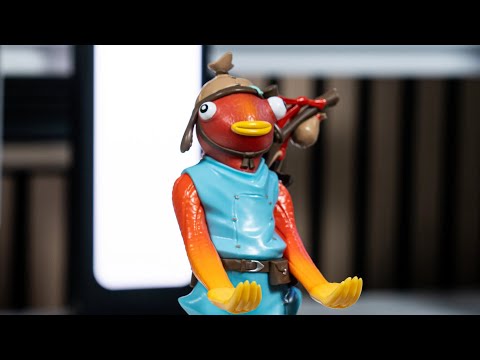 A Cable Guy figure of Fishstick from Fortnite standing upright with both hands extended to hold a controller or device. The character has its distinctive orange fish appearance, large cartoon eyes, and blue outfit, positioned on a sturdy display base.