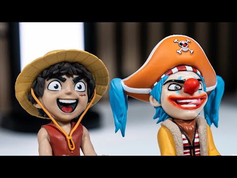 A rotating product showcase of two One Piece Mini Cable Guys stands. The Luffy mini stand is shown holding a phone-sized device with both hands, wearing his signature straw hat and red vest. The Buggy mini stand appears beside him, posed with outstretched hands and dressed in his clown-themed pirate outfit. The video highlights their small scale, painted details, and how they support small electronics on a desk.